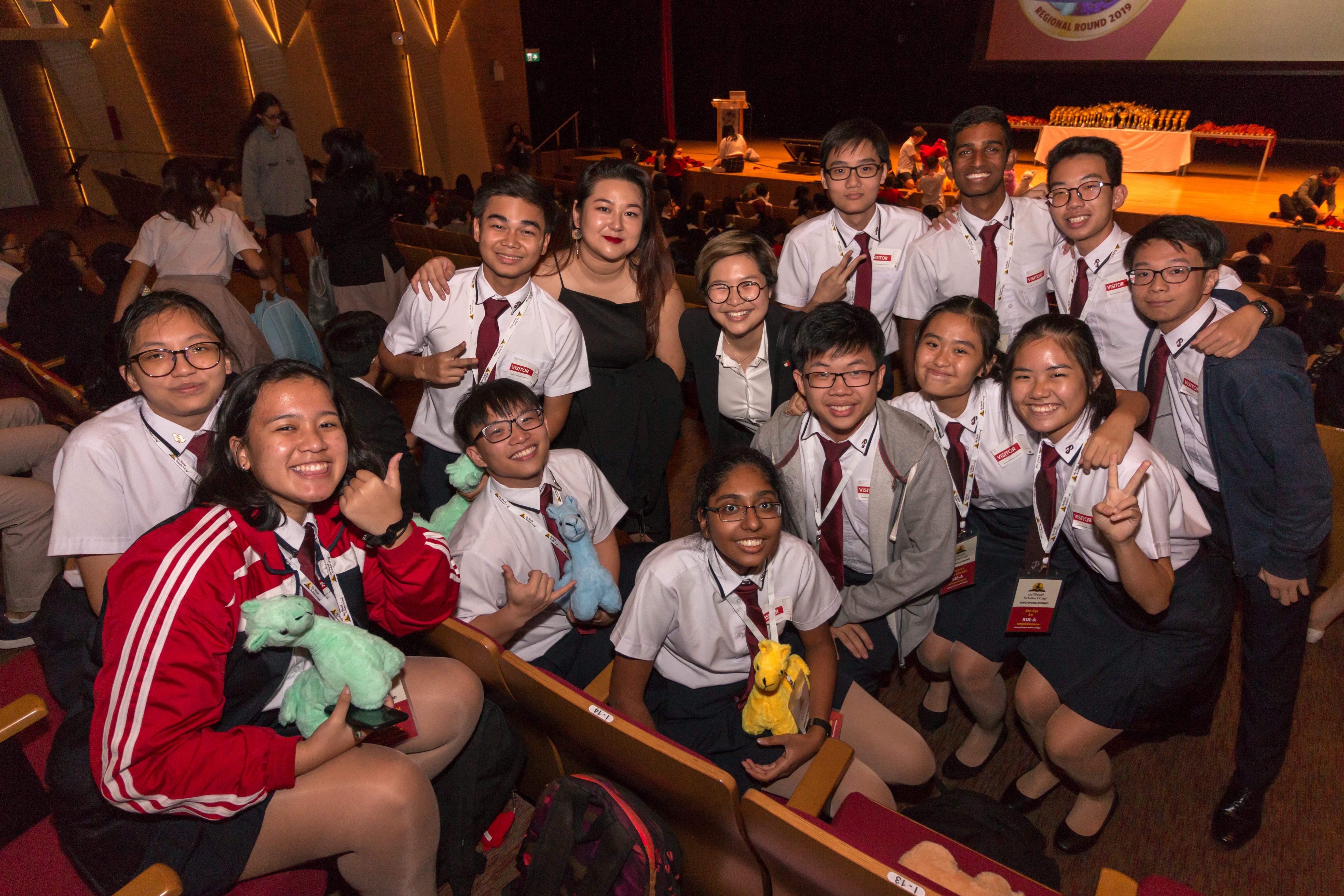 Group of students in school uniforms posing in a theater, smiling and holding plush toys.